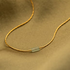 Close up of a dainty gold necklace featuring a small horizontal jade bar pendant, displayed on a soft fabric background.
