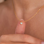 Gold necklace with a delicate chain featuring a small heart shaped opal pendant, held between fingers for detail.
