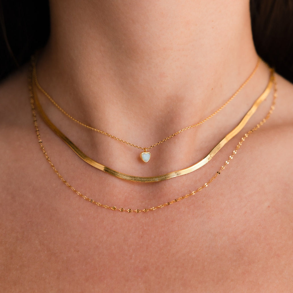 A person wearing three layered gold necklaces, including a charm necklace and the Tiny Opal Heart Necklace with a small heart-shaped pendant.