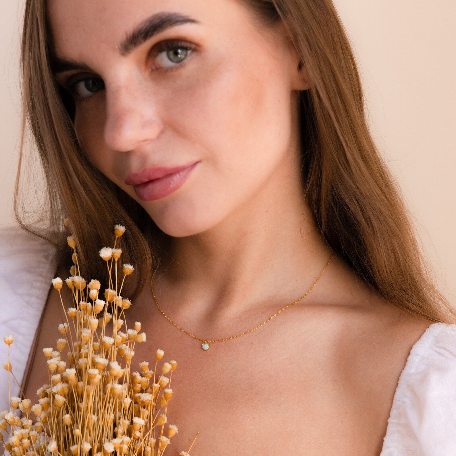 Woman with long brown hair holds dried flowers, wearing a white top and the Tiny Opal Heart Necklace, smiling softly.