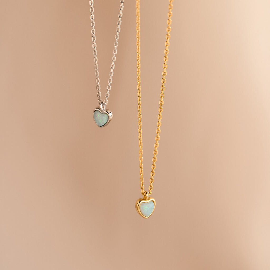 Two heart-shaped necklaces—one silver, one gold—with light blue stones rest on a beige background. Add the Tiny Opal Heart Necklace to your collection for a touch of charm.