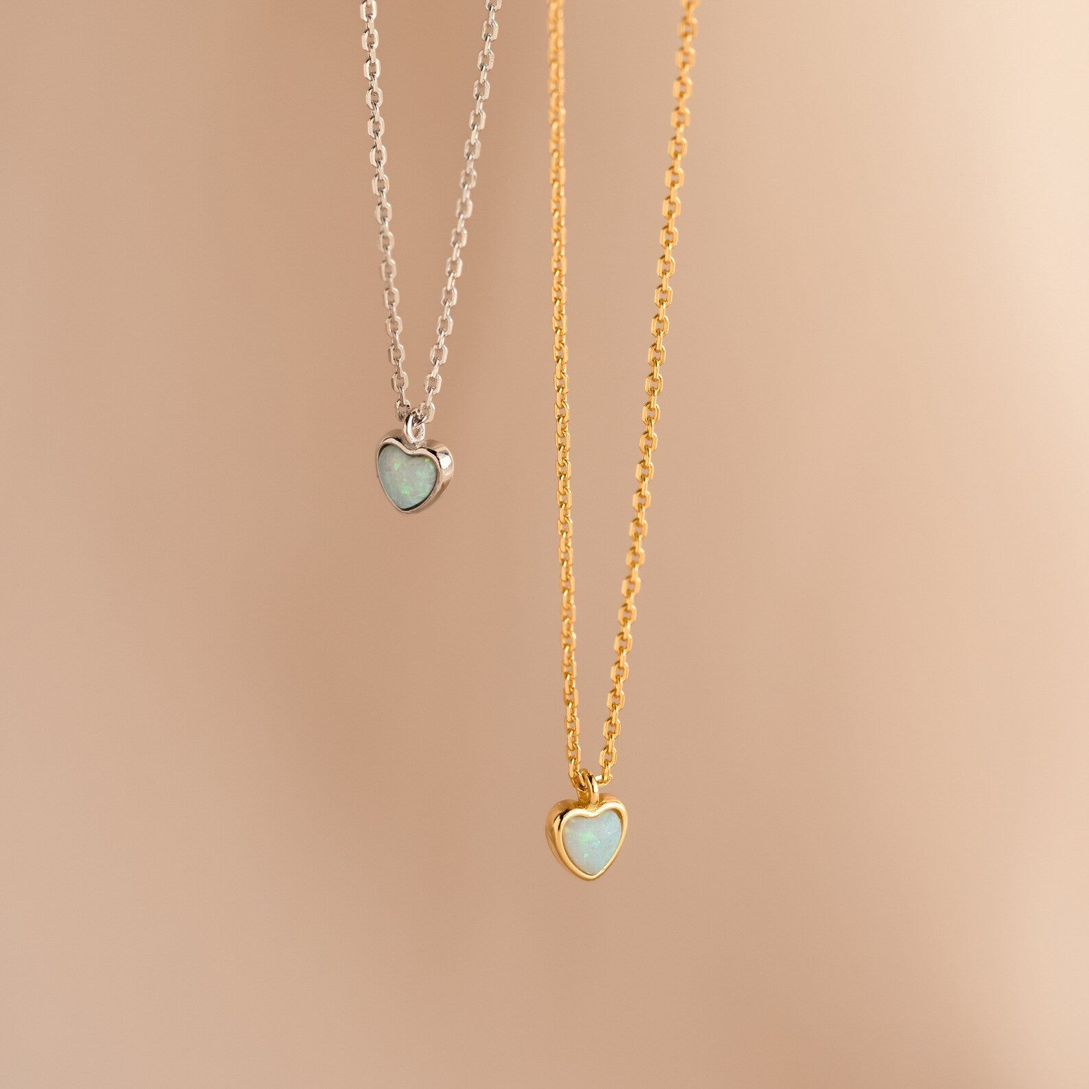 Two heart-shaped necklaces—one silver, one gold—with light blue stones rest on a beige background. Add the Tiny Opal Heart Necklace to your collection for a touch of charm.