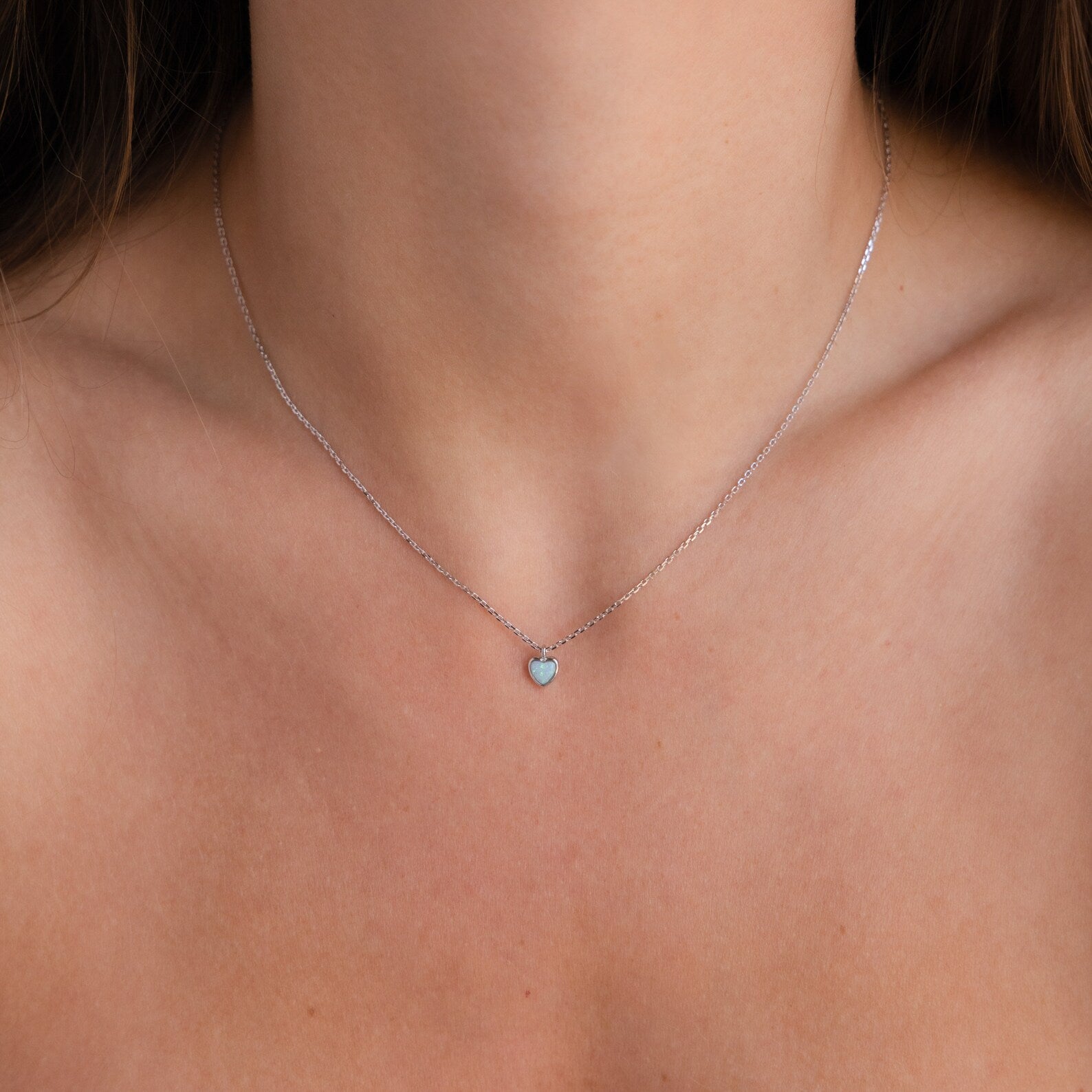 The Tiny Opal Heart Necklace, a delicate silver chain with a small opal heart pendant, rests gracefully on a woman's neck.
