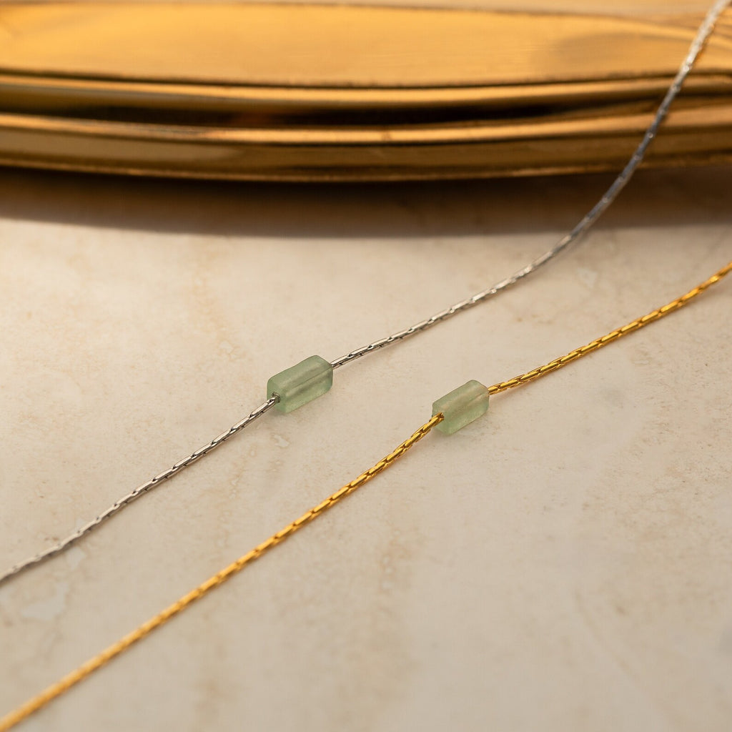 Two delicate chains, one silver and one gold, each featuring a green bead, capture the refined charm of the Dainty Aventurine Bar Necklace as they rest elegantly on a beige surface.