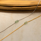 Two delicate chains, one silver and one gold, each featuring a green bead, capture the refined charm of the Dainty Aventurine Bar Necklace as they rest elegantly on a beige surface.