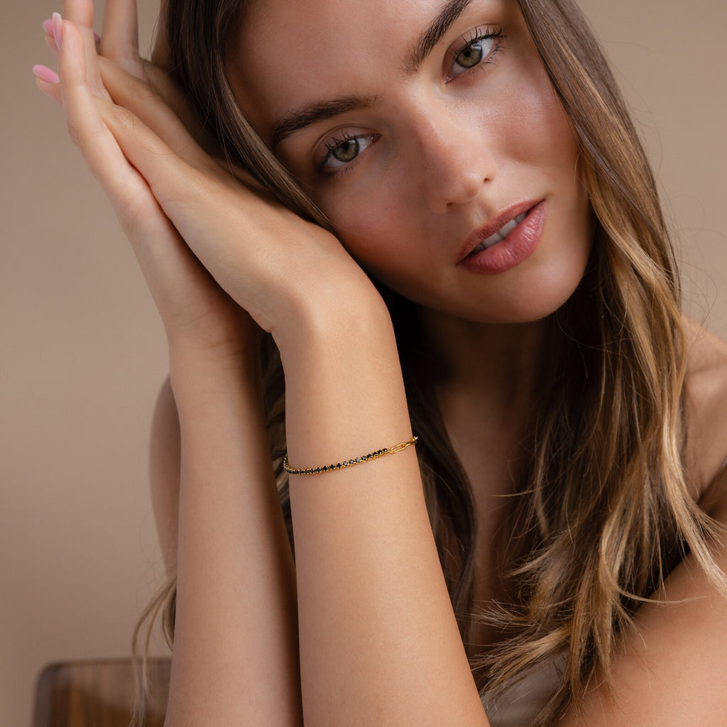A woman with long brown hair leans her head on her hands, showcasing the elegant Black Diamond Tennis Bracelet—a stunning statement piece—set against a beige background.