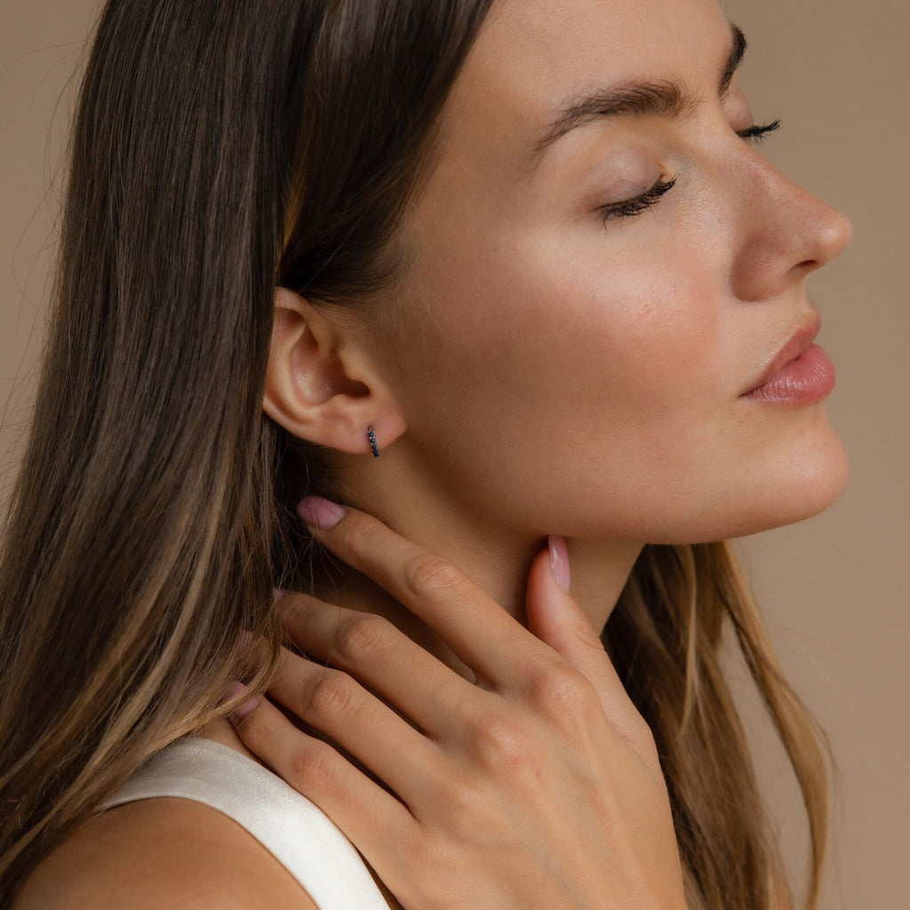 Woman with long hair and closed eyes touches her neck, wearing Indigo Blue Opal Huggies against a beige background.