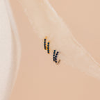 The Indigo Blue Opal Huggies: small gold and silver hoop earrings with deep blue stones, shown on a soft cream fabric for a minimalist look.