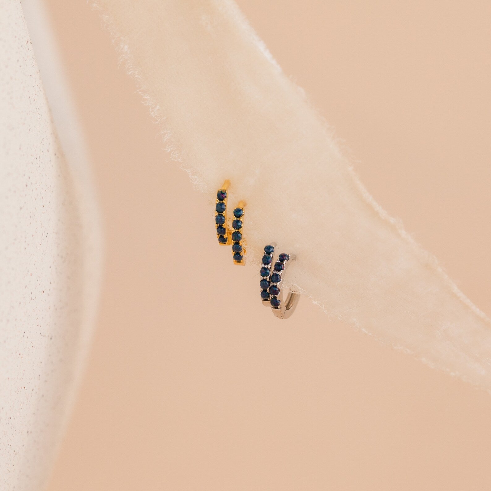 The Indigo Blue Opal Huggies: small gold and silver hoop earrings with deep blue stones, shown on a soft cream fabric for a minimalist look.