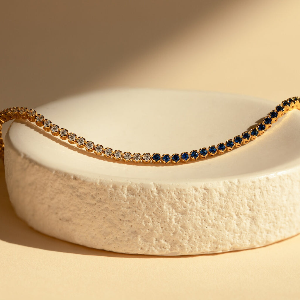 The Blue Diamond Tennis Bracelet, featuring alternating blue and clear stones, is displayed on a textured white dish—a minimalist and elegant accessory that makes the perfect "something blue" for special occasions.