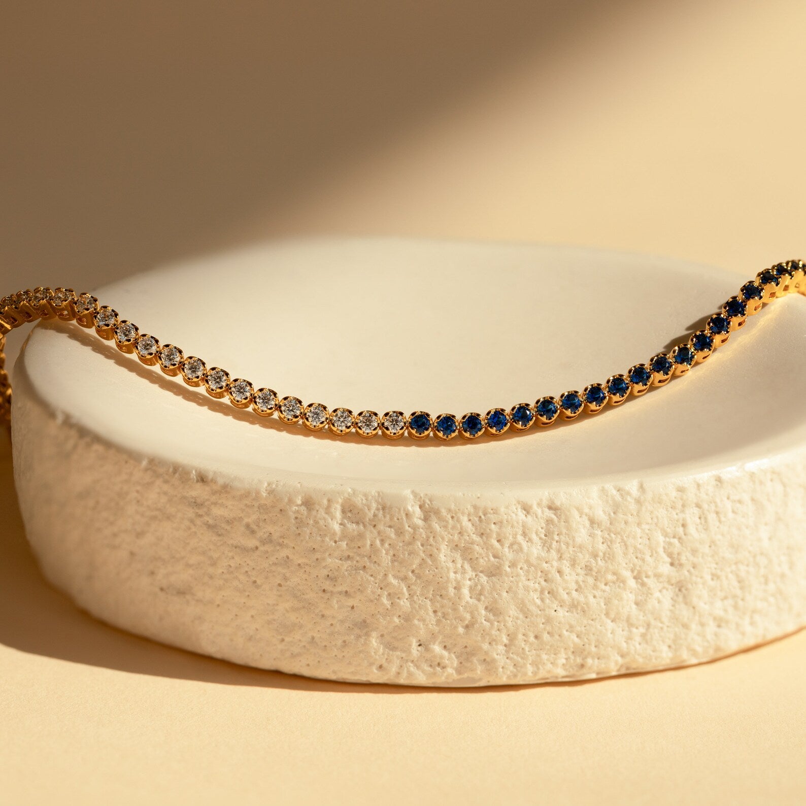 The Blue Diamond Tennis Bracelet, featuring alternating blue and clear stones, is displayed on a textured white dish—a minimalist and elegant accessory that makes the perfect "something blue" for special occasions.