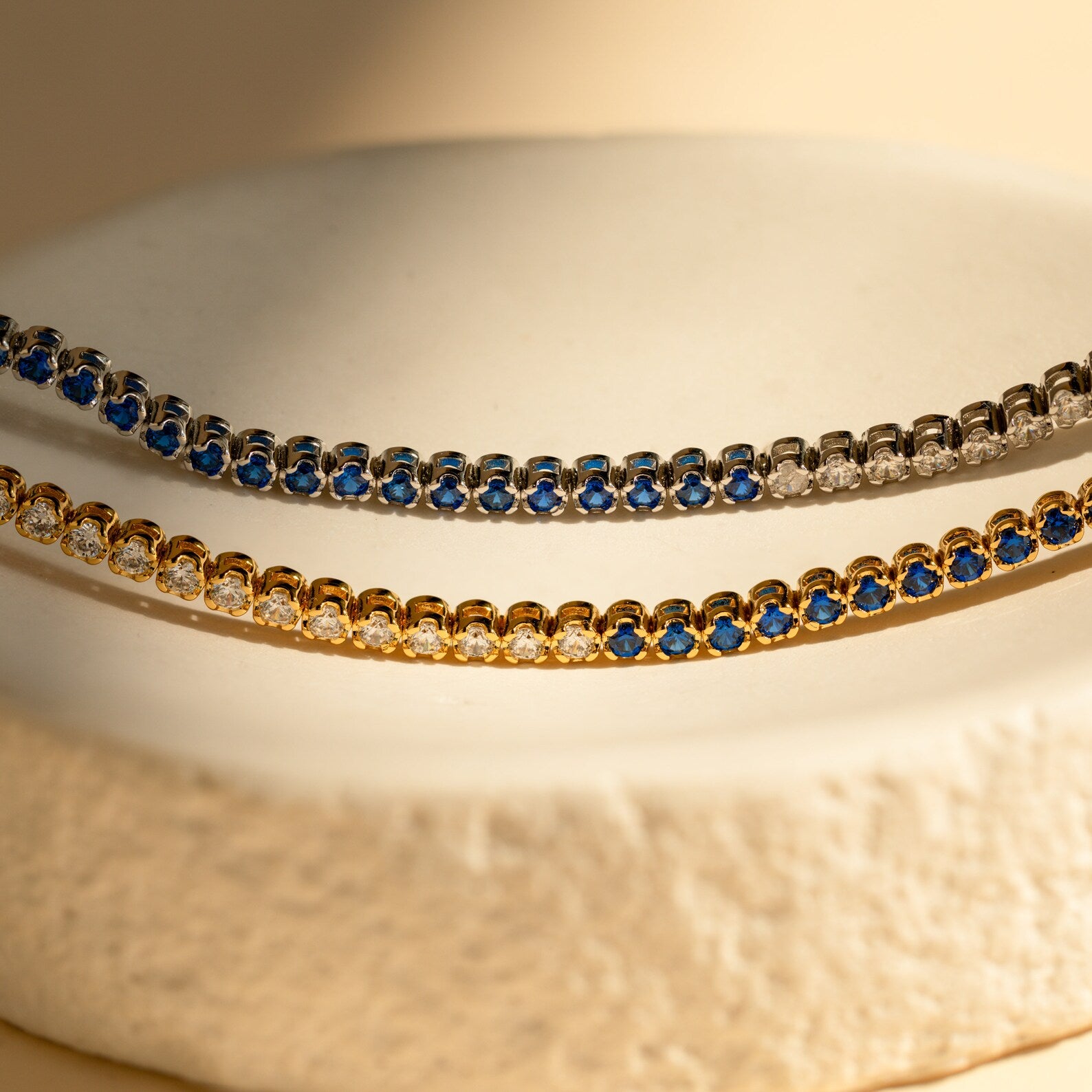 The Blue Diamond Tennis Bracelet features alternating blue and clear stones set in classic silver, making it a minimalist accessory and the perfect “something blue” for your look, elegantly displayed on a textured surface.