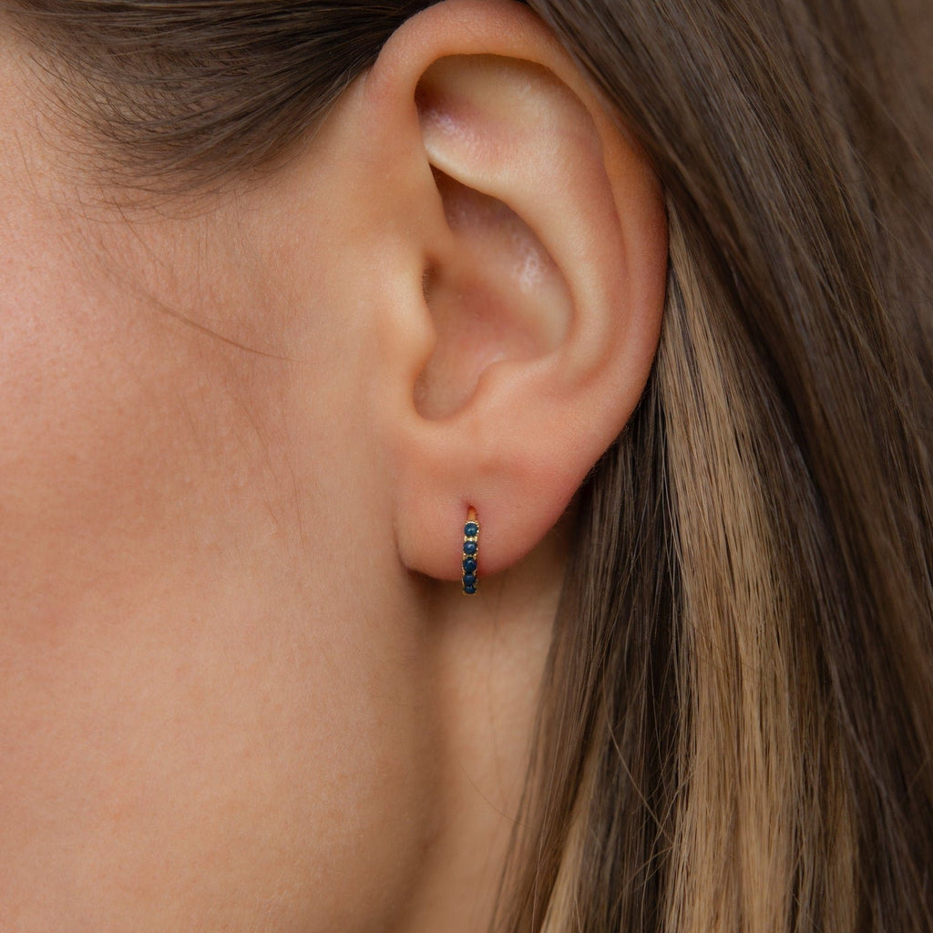 Close-up of a woman's ear wearing Indigo Blue Opal Huggies—minimalist gold hoop earrings accented with indigo blue opal stones.