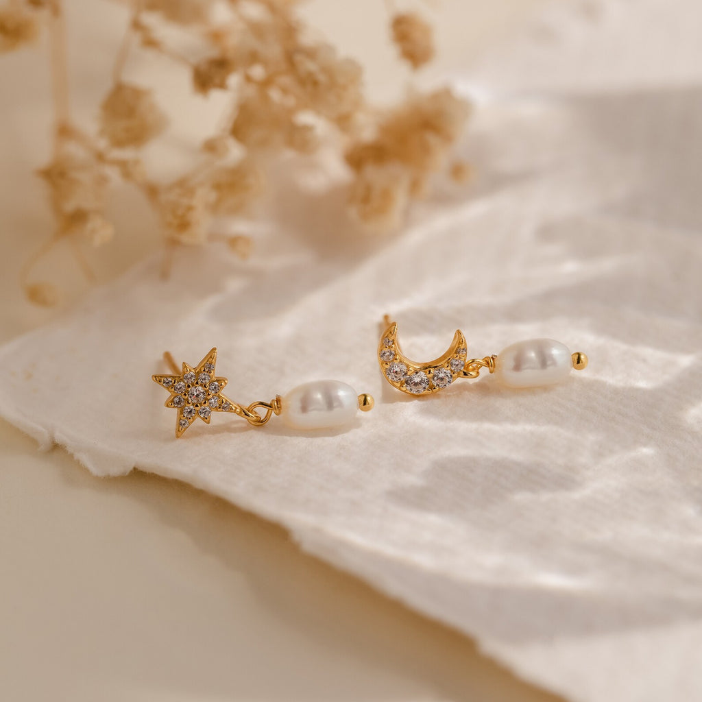 The Moon & Star Pearl Earrings, featuring delicate pearl charms, are displayed on white fabric with dried flowers in the background.