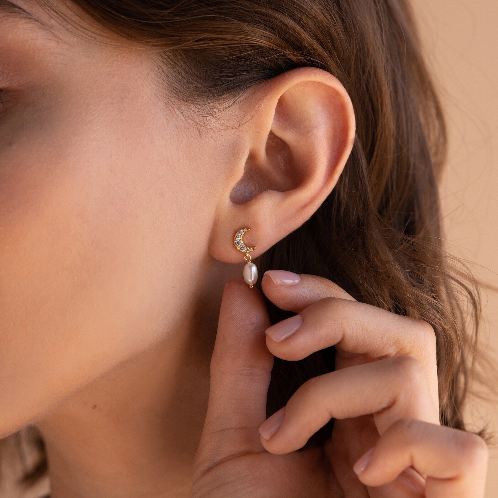 A close-up of a woman's ear wearing Moon & Star Pearl Earrings, featuring a gold crescent moon, pearl charm, and diamond gemstone stud, as her hand gently touches the earring.