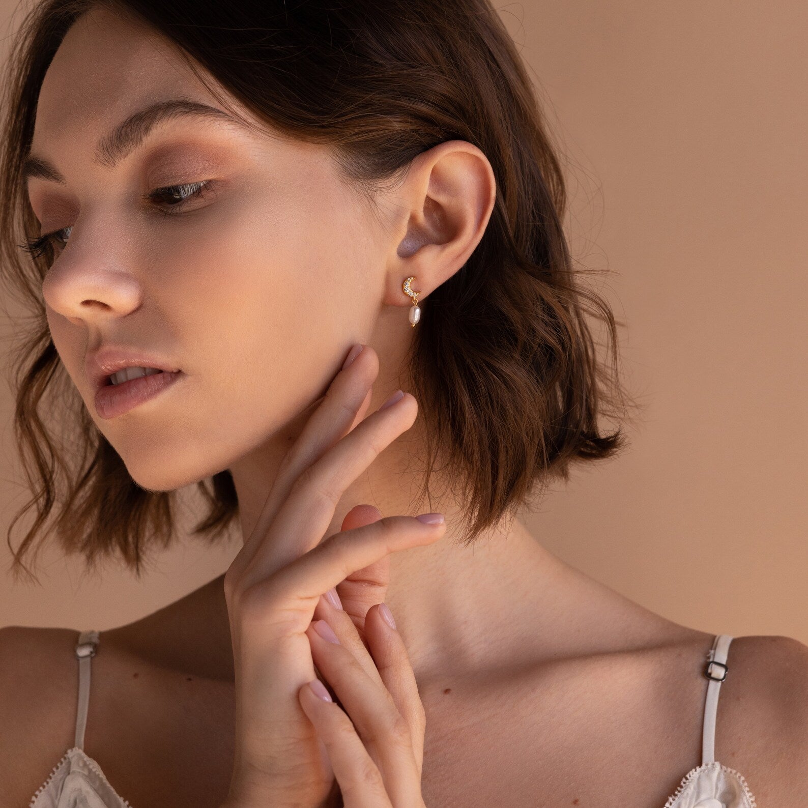 A woman with short brown hair wears a white top and Moon & Star Pearl Earrings, posing with her hand near her face.