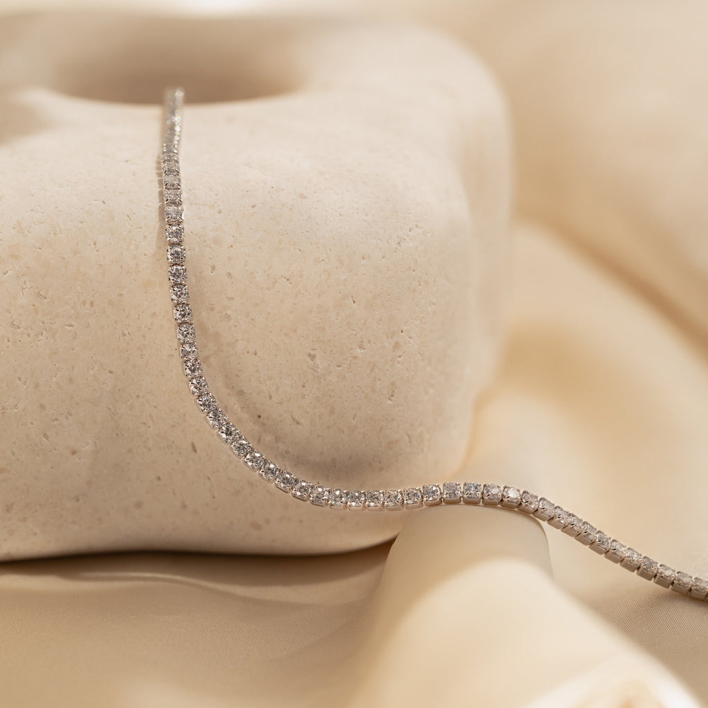 The Dainty Diamond Tennis Necklace & Bracelet Set in White Gold is elegantly displayed draped over a smooth stone and beige satin fabric.