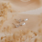Moon & Star Pearl Earrings, featuring delicate pearl charms and diamond gemstone studs, are showcased on soft beige fabric with blurred floral accents.
