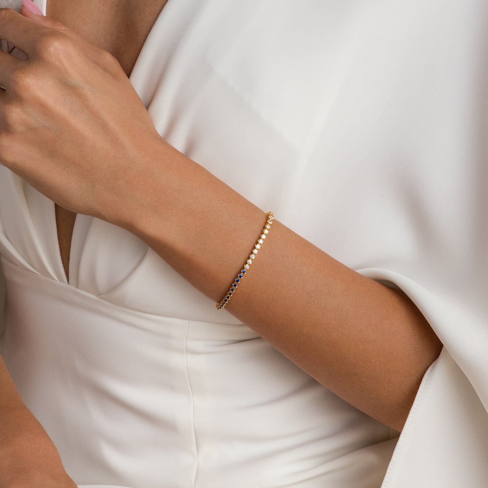 Woman in a white dress wears the Blue Diamond Tennis Bracelet, a minimalist gold accessory, while holding a cup near her chest.
