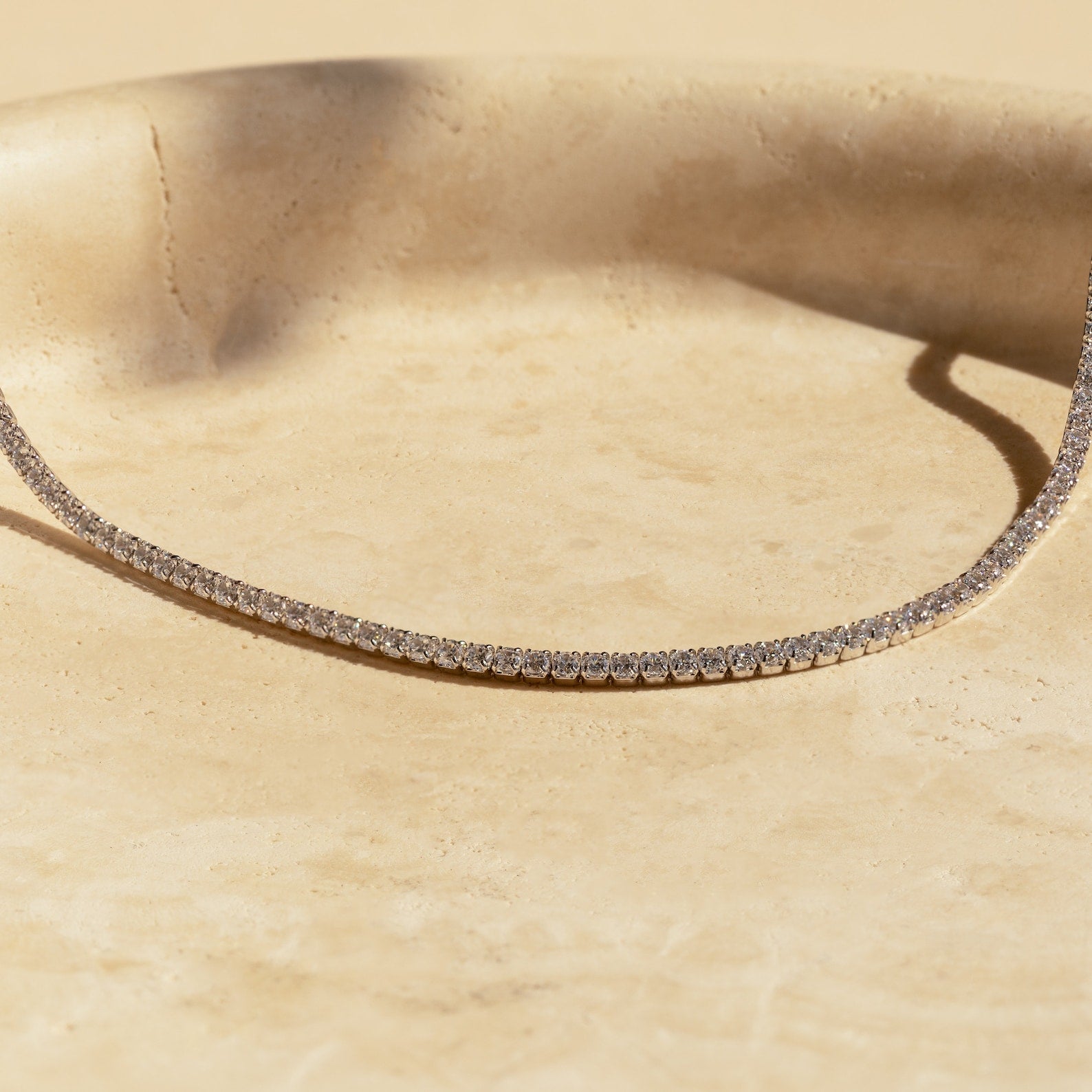 White gold diamond tennis choker necklace arranged in a graceful curve on a light stone surface, with round-cut diamonds sparkling under natural sunlight.