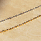 The Tennis Necklace Set, featuring one silver and one gold necklace, is displayed on a beige textured surface—perfect for layering your jewelry style.