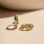 A pair of gold huggie earrings set on a soft cream surface, each featuring three icy blue aquamarine gemstones. The design showcases a vertically stacked round, princess-cut, and oval aquamarine stone, delicately prong-set in a high-polished gold setting. The light-catching facets of the stones reflect soft blue tones, creating an elegant contrast against the warm gold