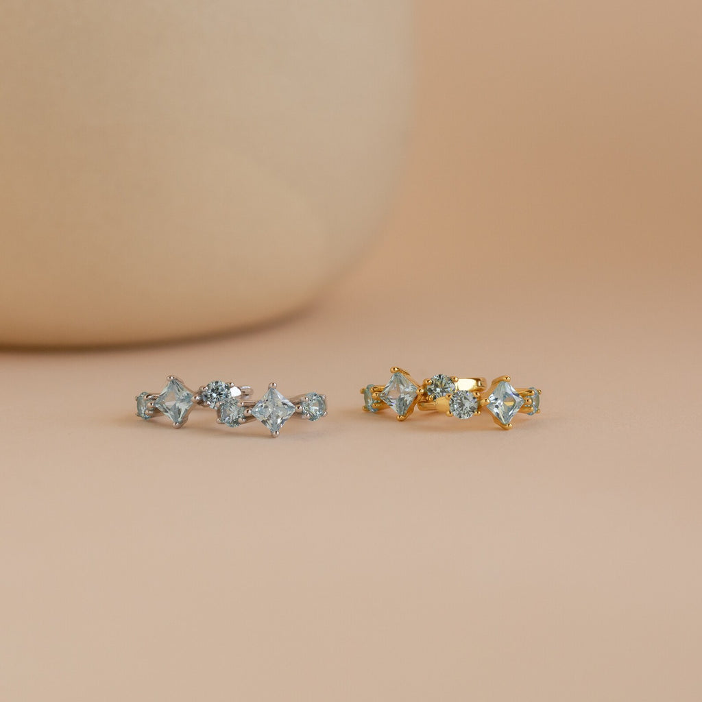 Side-by-side display of both gold and silver Aquamarine Diamond Huggies on a soft peach backdrop.