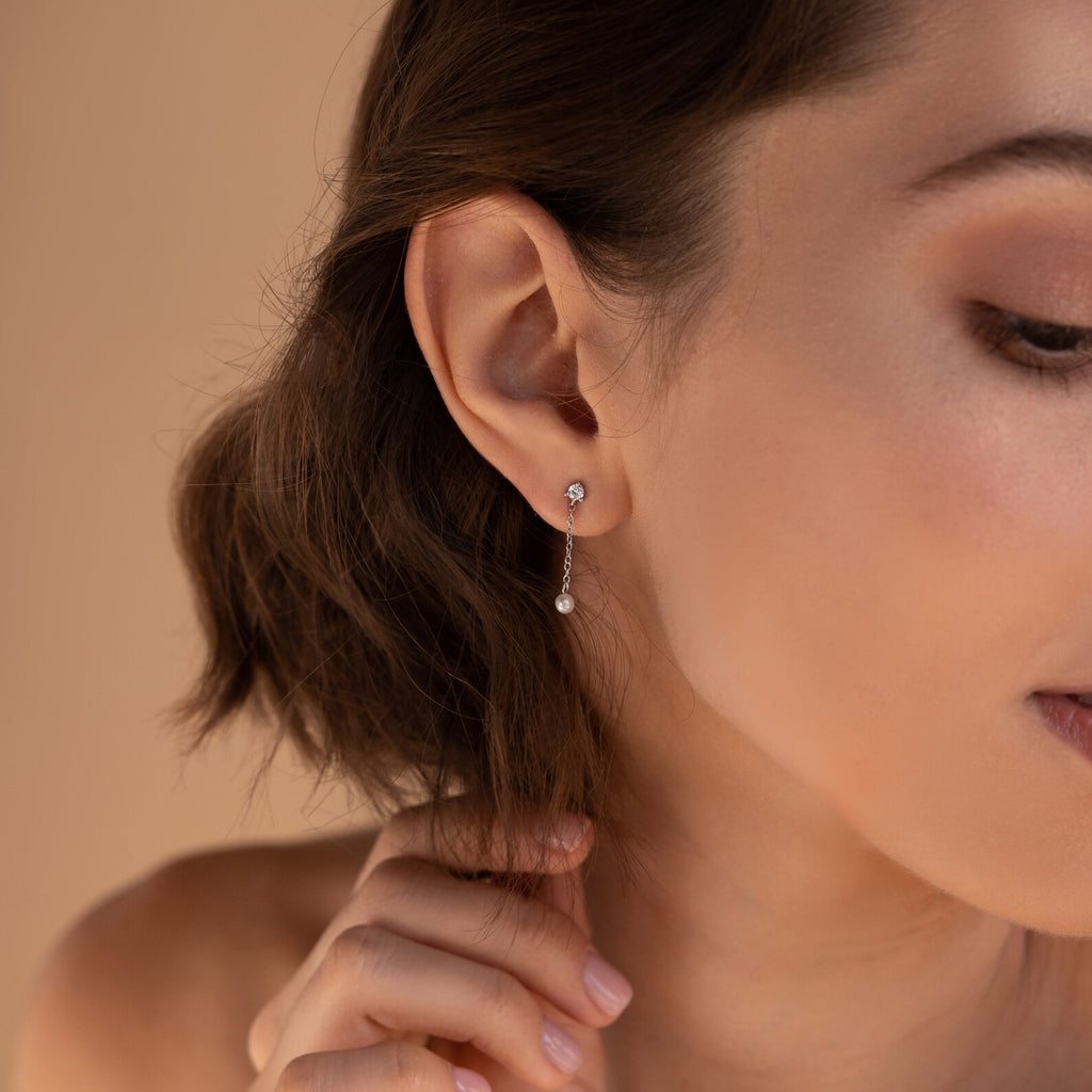 A woman touches her hair, showcasing elegant Diamond Pearl Drop Earrings in her pierced ear against a neutral background.