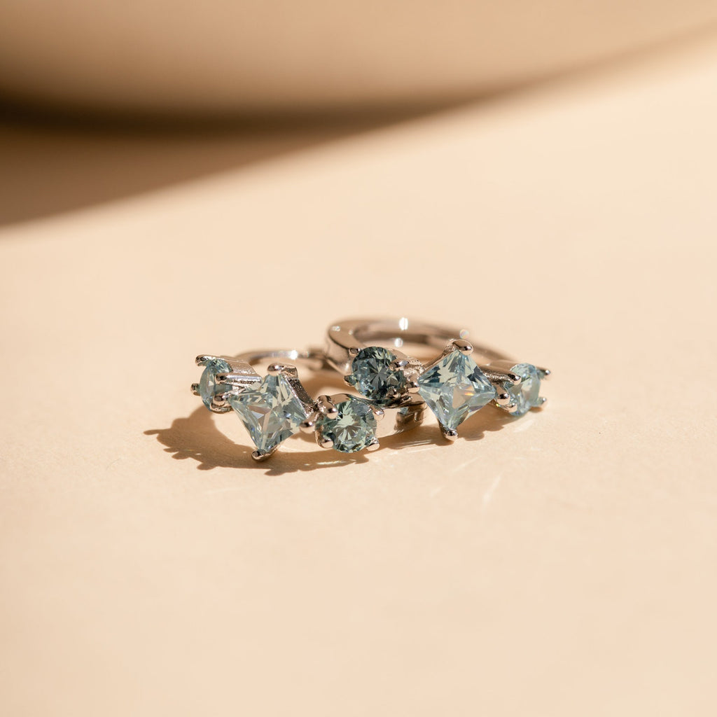 Close-up view of the silver Aquamarine Diamond Huggies lying on a peach-toned surface, with their gemstones shimmering under light.
