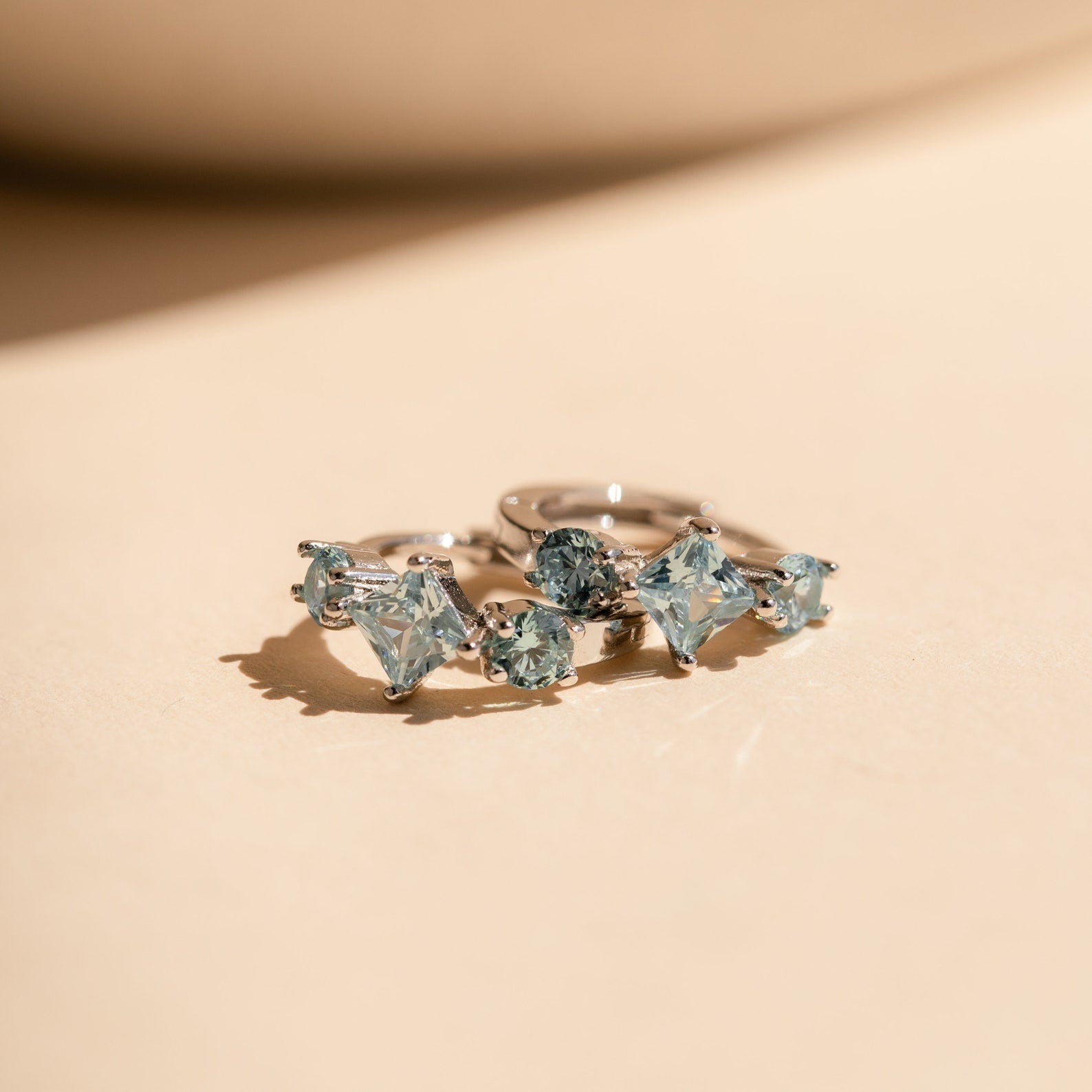 Close-up view of the silver Aquamarine Diamond Huggies lying on a peach-toned surface, with their gemstones shimmering under light.