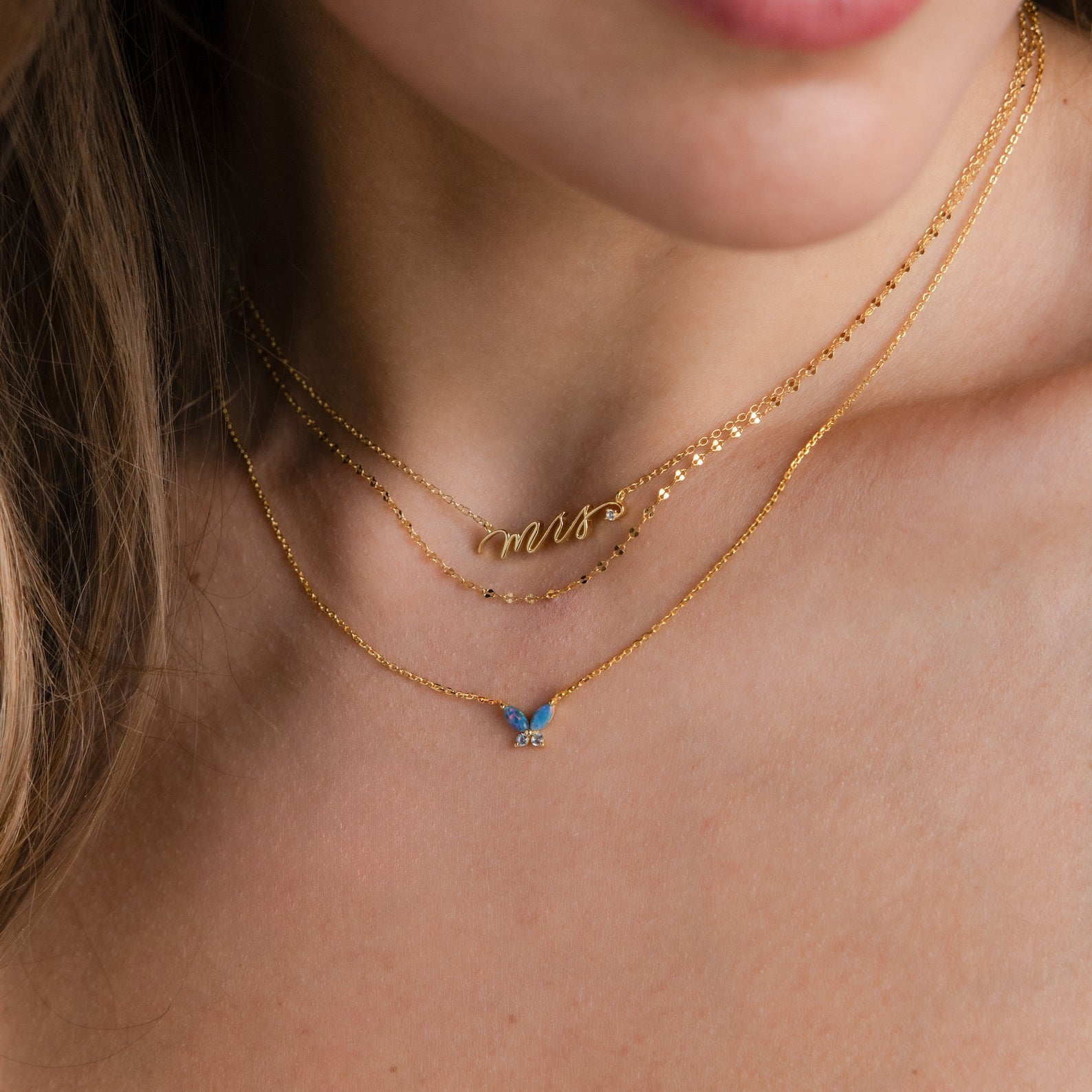 Close-up of a woman wearing layered gold necklaces, featuring a dainty "mrs" piece and the Blue Opal Butterfly Necklace with a shimmering blue butterfly charm.