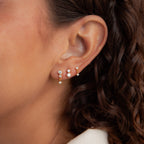 A close-up of a woman's ear shows the Triple Diamond Studs Set and stacking gold earrings; her curly brown hair is visible.