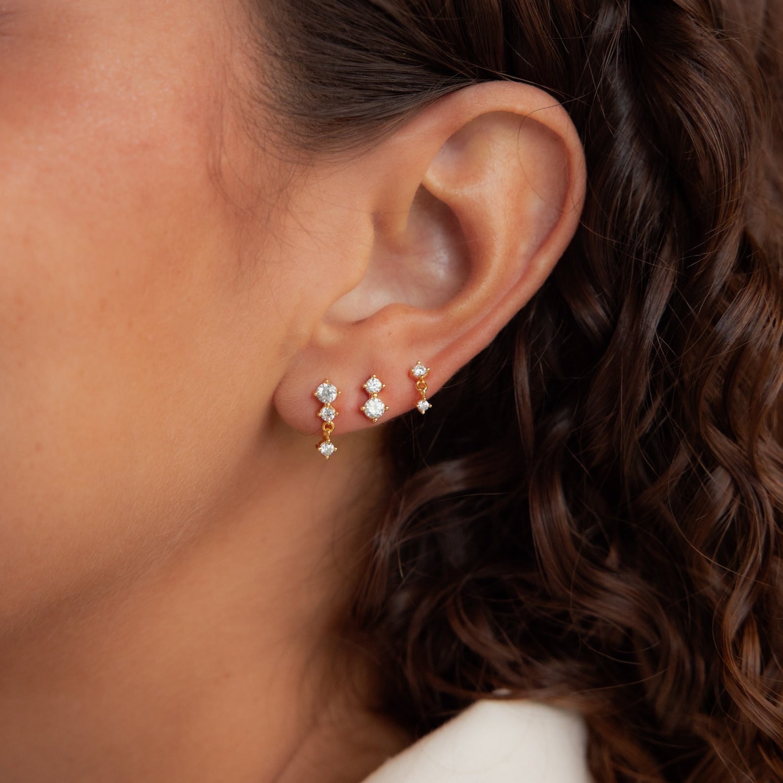 A close-up of a woman's ear shows the Triple Diamond Studs Set and stacking gold earrings; her curly brown hair is visible.
