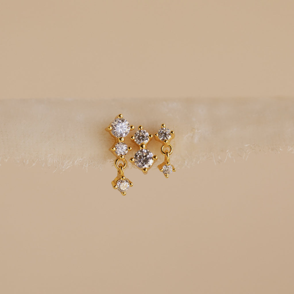 The Triple Diamond Studs Set features gold stacking earrings, each with three clear gemstones reminiscent of diamond drops, showcased on a soft neutral background.
