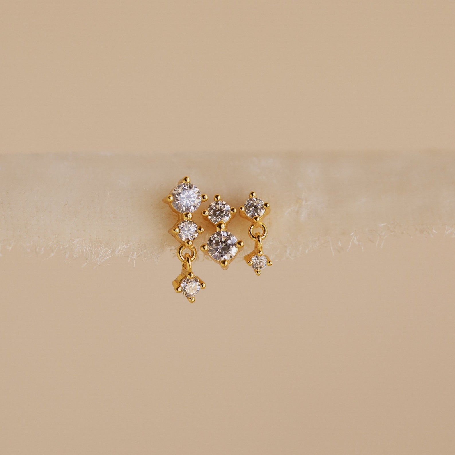 The Triple Diamond Studs Set features gold stacking earrings, each with three clear gemstones reminiscent of diamond drops, showcased on a soft neutral background.