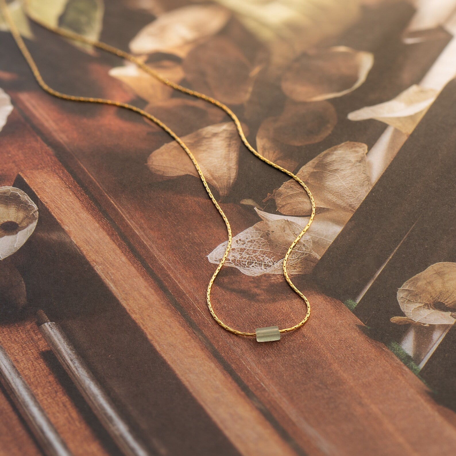 The Dainty Aventurine Bar Necklace, featuring a slim gold rectangular pendant, is displayed on a floral-patterned surface.