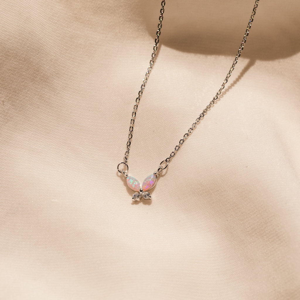 The Pink Opal Butterfly Necklace features a silver chain and an iridescent butterfly pendant, beautifully displayed on a soft beige fabric background.