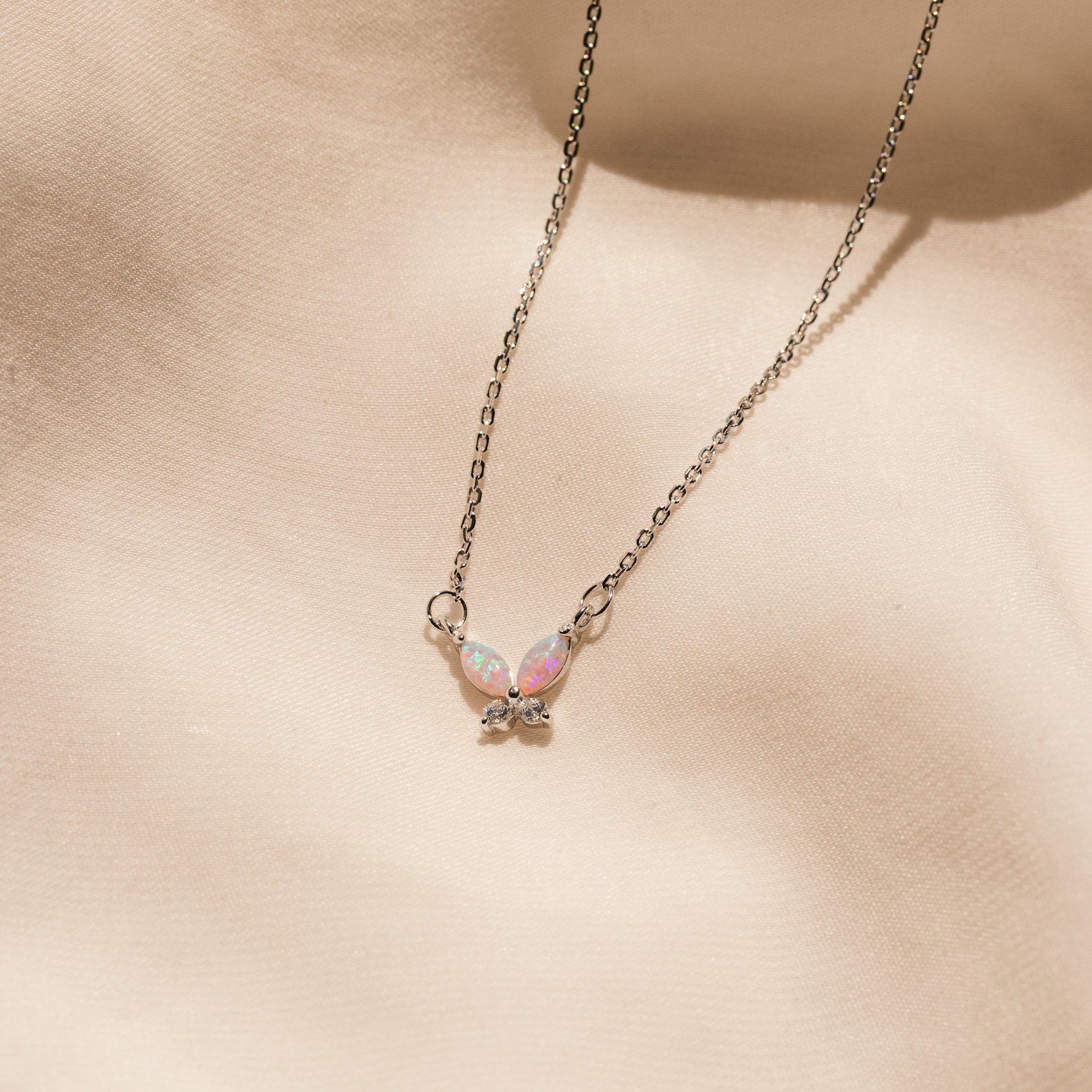 The Pink Opal Butterfly Necklace features a silver chain and an iridescent butterfly pendant, beautifully displayed on a soft beige fabric background.