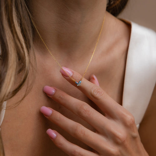 A woman with pink-manicured nails gently touches her Blue Opal Butterfly Necklace, showcasing its delicate butterfly pendant.
