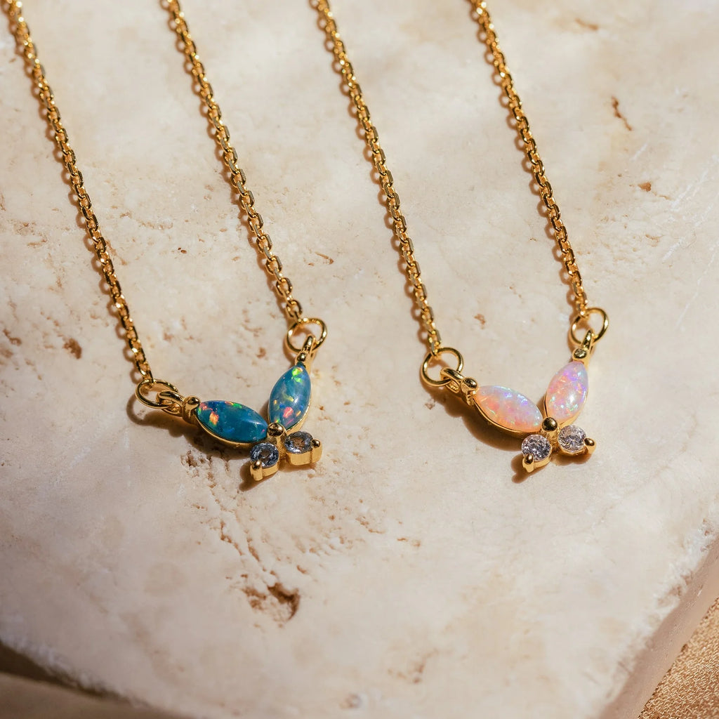 The Opal Butterfly Necklace Set features two gold chain necklaces with butterfly-shaped pendants—one pink opal and one blue opal—both accented with sparkling gems, elegantly displayed on stone.