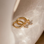 Close-up of a pair of small gold huggie hoop earrings, each featuring a dangling four-petal diamond flower charm. The hoops have a smooth, polished finish, while the flower charms are encrusted with sparkling clear stones.
