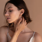 A woman gazes downward with one hand gently touching her face. She wears a gold Diamond Flower Huggie and a matching bracelet and ring, styled together to complete a cohesive, delicate jewelry look