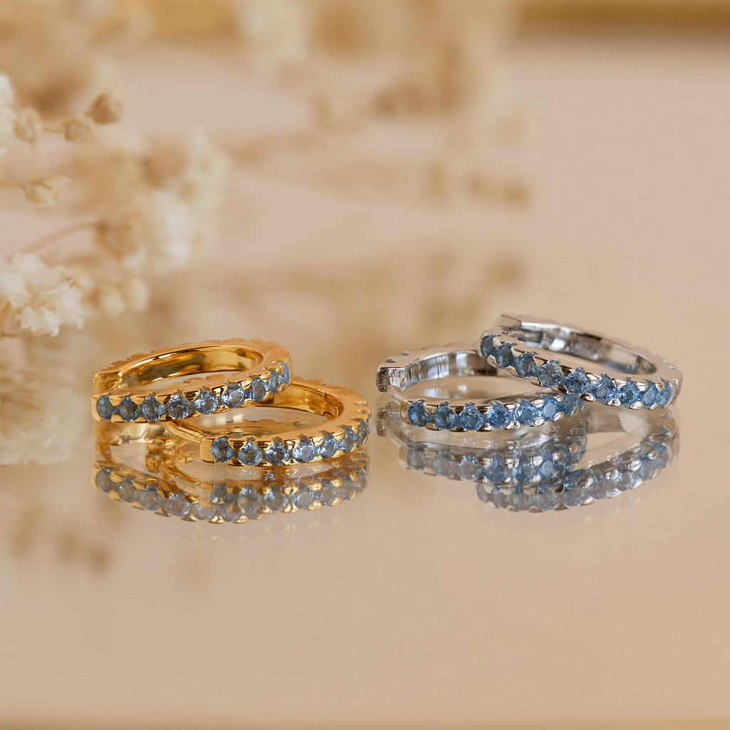 Two pairs of gemstone hoop earrings—one gold, one silver—are displayed on a reflective surface, perfect for stacking or pairing with Aquamarine Pave Huggies for an elegant look.