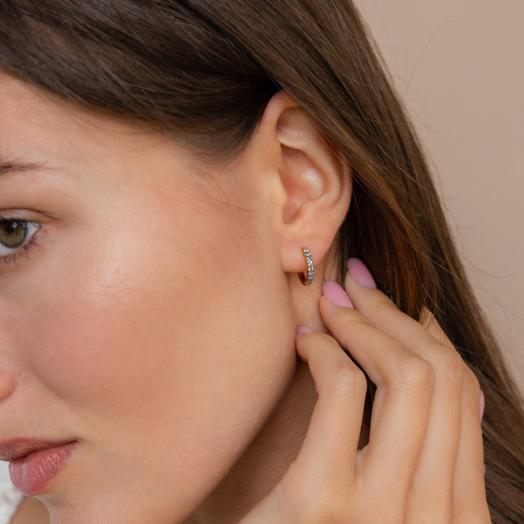 Woman touching her ear, wearing Aquamarine Pave Huggies—small gold hoop earrings with aquamarine pave stones—styled in an elegant earring stack, close-up side profile.
