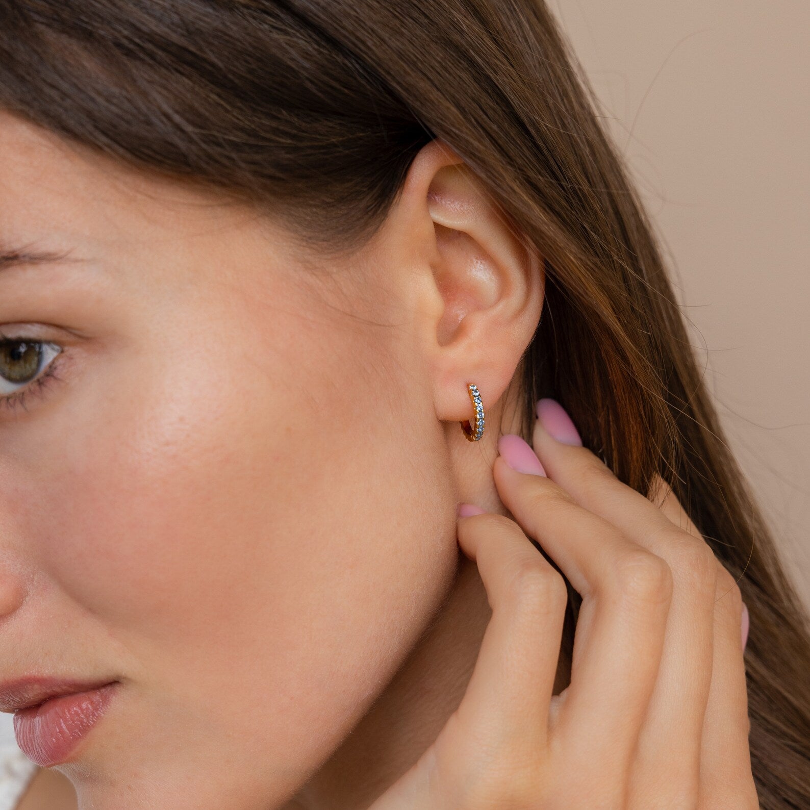 Woman touching her ear, wearing Aquamarine Pave Huggies—small gold hoop earrings with aquamarine pave stones—styled in an elegant earring stack, close-up side profile.