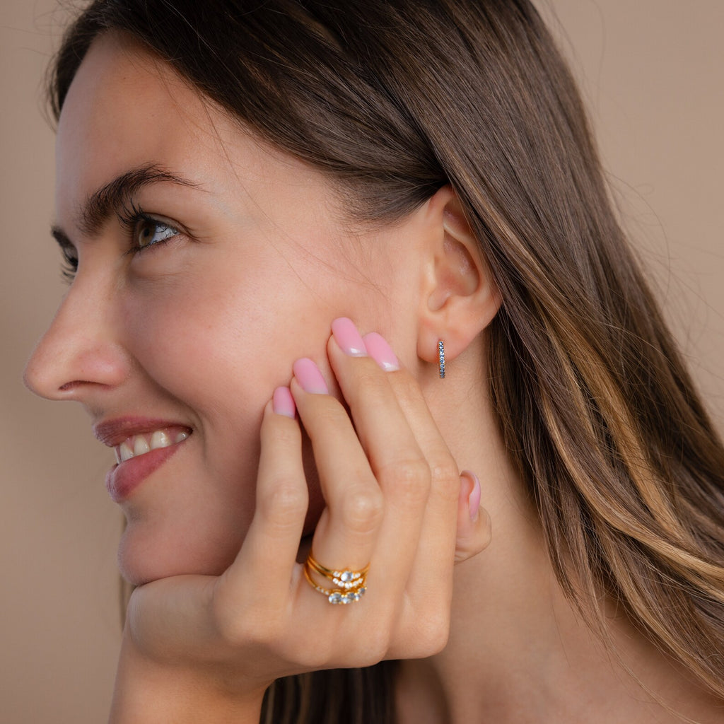 Woman with long brown hair smiles, showing pink nails, a ring, and a stylish earring stack featuring Aquamarine Pave Huggies.