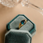 The London Blue Signet Ring features a vibrant London Blue Topaz set in a gold band, presented in a green velvet jewelry box with a shimmering glass object in the background.