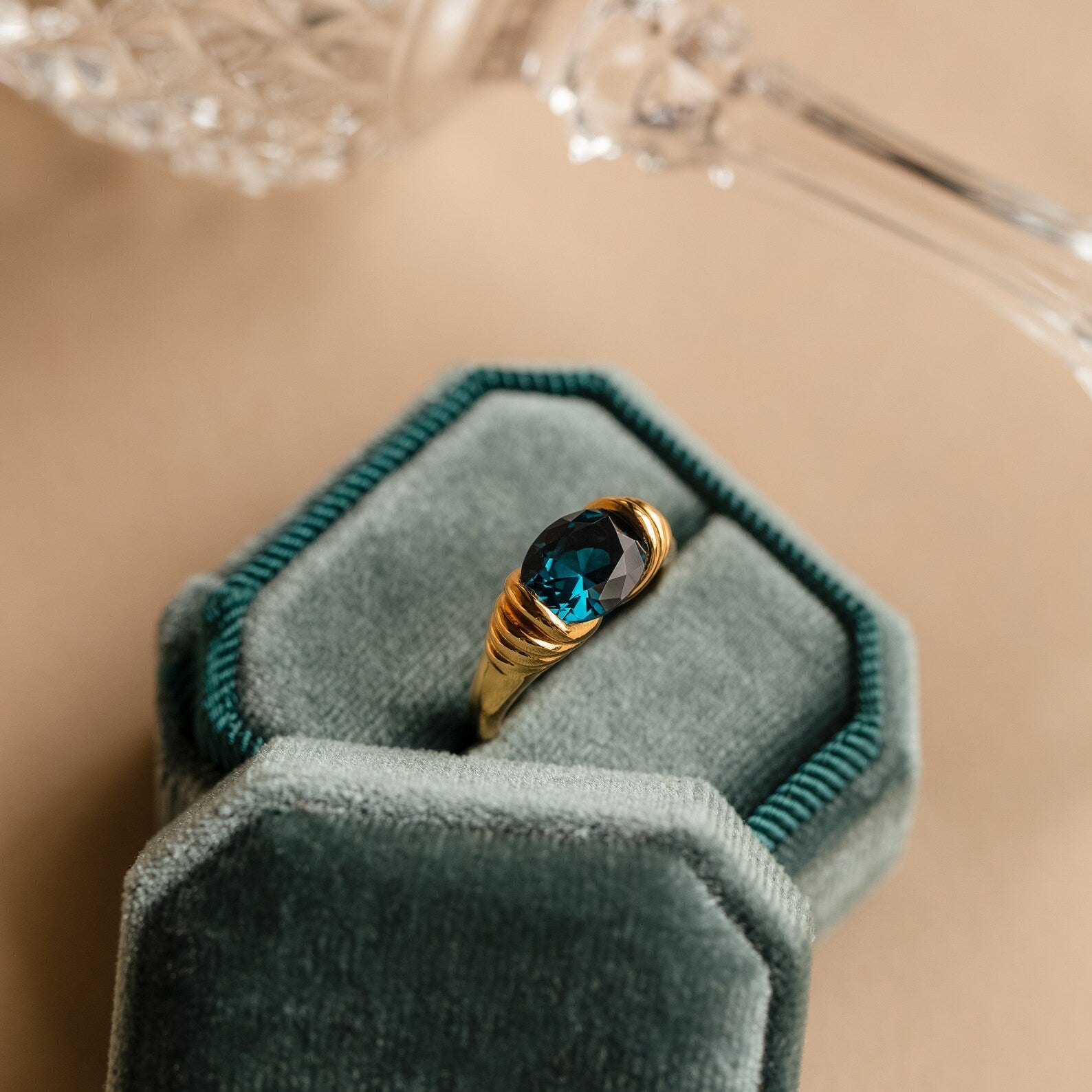 The London Blue Signet Ring features a vibrant London Blue Topaz set in a gold band, presented in a green velvet jewelry box with a shimmering glass object in the background.