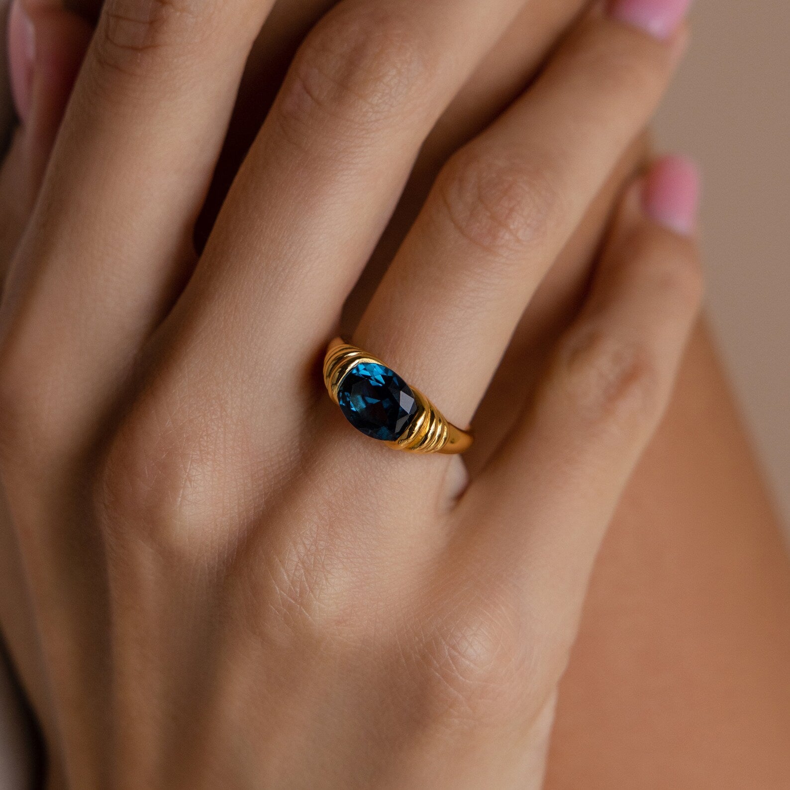 A hand with pink nails wears the London Blue Signet Ring—a gold band featuring an oval London Blue Topaz gemstone—on the ring finger.