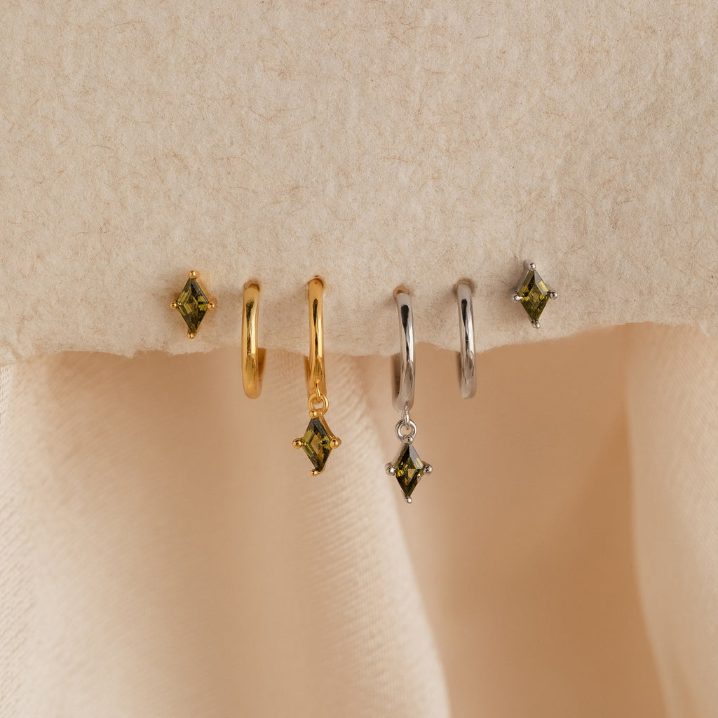 The Peridot Earrings Set in 18K Gold features two pairs of gold and silver hoop stacking earrings with green square gemstones, displayed on a beige textured surface.